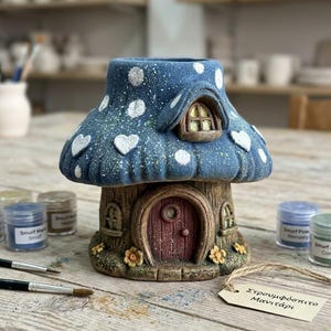 Può includere: Una casa in ceramica a forma di fungo con un cappello blu testurizzato decorato con pois e cuori bianchi. La base ricorda un tronco d'albero con una porta ad arco marrone e piccole finestre. Un'etichetta riporta il testo "Στρουμφόσπιτο Μανιτάρι".