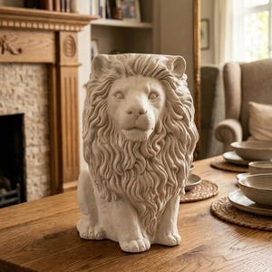 May include: A cream-colored ceramic lion sculpture featuring a detailed, flowing mane and a seated posture. The lion has a neutral expression with visible facial features, including eyes, a nose, and a mouth, rendered in a matte, stone-like finish.