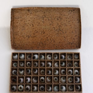 Puede incluir: Una caja rectangular de cartón con tapa marrón moteada y base compartimentada que contiene cuarenta y cinco prótesis oculares de vidrio. Cada prótesis presenta una esclerótica blanca y un iris oscuro, dispuestas en cinco filas.