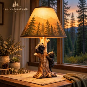 May include: A table lamp featuring a base sculpted as a tree trunk with two black bear cubs climbing it. The conical lampshade displays a printed landscape of evergreen trees and mountains in warm orange and brown tones. The text TimberAuraCrafts is visible.