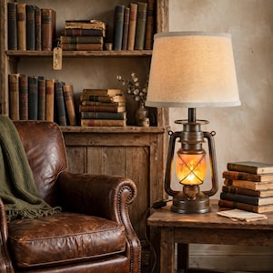 May include: A table lamp featuring a dark bronze metal lantern base with a glowing amber glass center and a tapered beige fabric lampshade. The lamp sits on a wooden surface next to a stack of vintage books, with a leather armchair nearby.