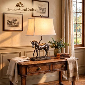 May include: A table lamp featuring a bronze-colored horse sculpture as the base, topped with a rectangular off-white fabric lampshade. The lamp sits on a rectangular base and includes a small horseshoe finial at the top of the shade frame.