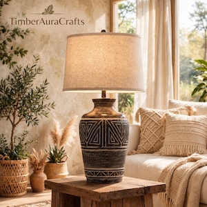 May include: A table lamp with a beige fabric drum shade and a dark brown, textured ceramic base featuring carved geometric patterns. The lamp sits on a wooden surface, displaying a cylindrical body with horizontal and triangular etched designs.