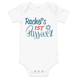 May include: White baby bodysuit with the text "Rachel's 1st Passover" in blue and red script. A blue Star of David is next to the word "1st". The bodysuit has short sleeves and poppers at the bottom.