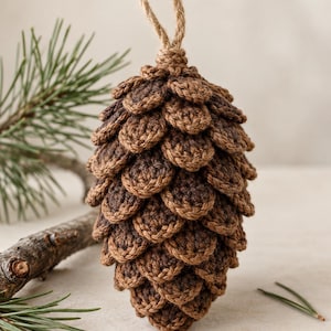 Può includere: Un ornamento a forma di pigna realizzato all'uncinetto in filo marrone, caratterizzato da squame stratificate e testurizzate e da un cappio in corda di juta naturale sulla parte superiore. L'oggetto artigianale imita una pigna vera.