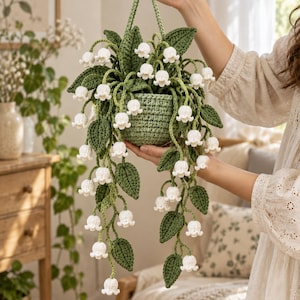 May include: A crocheted hanging plant featuring white lily of the valley flowers with bell-shaped blooms and dark green leaves. The plant is contained in a textured, light green crocheted pot and includes a matching green crocheted hanging loop.