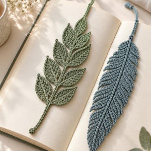 May include: Two handmade crochet bookmarks resting on an open book. One bookmark is a sage green branch with multiple leaves, and the other is a dusty blue feather shape. Both items feature intricate yarn stitching and a small loop at the top.