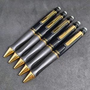 Op de afbeelding: Vijf zwart-gouden mechanische potloden diagonaal gerangschikt. Elk potlood heeft een gouden clip, een gouden punt, een getextureerd donkergrijs gripgedeelte en een witte gum aan de bovenkant. Het woord PhD is in goud op de houder gedrukt.