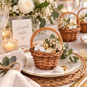 May include: A small, round, woven brown wicker basket with a handle, filled with white and metallic gold almond candies, sprigs of white baby's breath flowers, and green eucalyptus leaves, resting on a white plate with a gold rim.