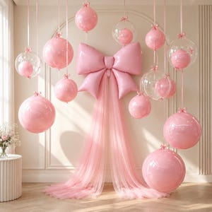 Peut inclure: Une décoration composée d'un grand nœud en aluminium rose avec de longs rubans en tulle, entouré de divers ballons roses et de ballons transparents contenant de petites sphères roses, le tout suspendu par des rubans fins.