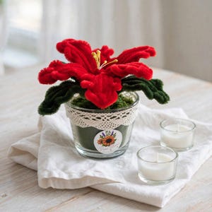 Pode incluir: Uma flor de lírio vermelha feita de limpadores de cachimbo com estames amarelos e folhas verde-escuras, num frasco de vidro transparente com rebordo de renda branca. Um autocolante circular com um girassol e o texto Pure Blooms está no vidro.