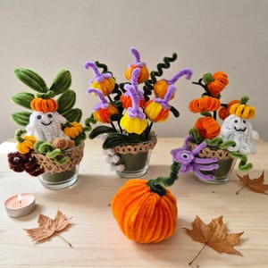 May include: Three small glass pots wrapped in burlap ribbon contain pipe cleaner crafts featuring orange pumpkins, green leaves, and white ghosts with black eyes and mouths. A single large orange pipe cleaner pumpkin sits in front.