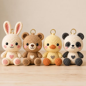 Può includere: Quattro portachiavi a forma di animale realizzati all'uncinetto, tra cui un coniglio color crema con dettagli rosa, un orso marrone, un'anatra gialla con il becco arancione e un panda bianco e grigio scuro. Ogni figura ha occhi neri e un cuore.