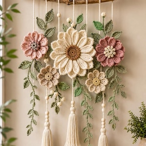 May include: A wall hanging featuring crocheted flowers in shades of cream, dusty pink, and beige, accented with green crocheted leaves. The floral arrangement is suspended from a natural wooden branch by cream cords, finished with three long tassels.