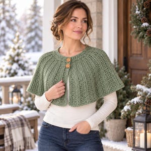 Könnte beinhalten: Ein salbeigrünes Häkel-Capelet mit gerippter Textur und zwei runden Holzknöpfen am Halsausschnitt. Das Kleidungsstück wird über einem langärmeligen weißen Oberteil und einer dunkelblauen Jeans getragen, was einen Lagenlook mit Rundhalsausschnitt ergibt.