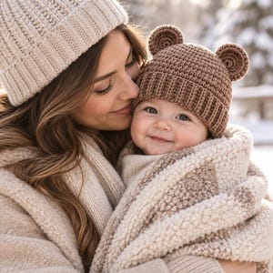 May include: A beige knit beanie with a pom-pom and a brown crochet beanie featuring two small rounded bear ears on top. The items are paired with a textured beige and brown fleece blanket, showcasing a soft, thick material and neutral colour palette.