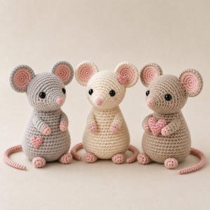 Op de afbeelding: Drie gehaakte muispoppen met ronde oren, zwarte kraaloogjes en roze accenten. De linker muis is grijs met een roze hartje, de middelste is crème en de rechter is beige en houdt een roze hartje vast. Elke muis heeft een lange staart en kleine roze pootjes.