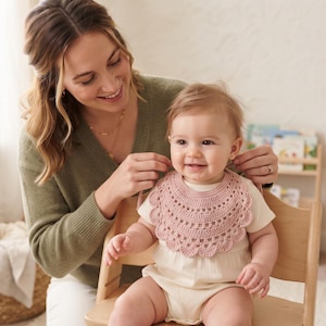 Può includere: Una bavaglino per neonati all'uncinetto color rosa polvere con bordo smerlato e design circolare. Il bavaglino è indossato sopra un body a maniche corte color crema, mostrando complessi punti di filato e un aspetto morbido e strutturato.