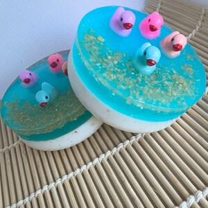 Puede incluir: Dos pastillas de jabón redondas con un diseño en capas, con una base blanca opaca y una capa superior azul translúcida. La sección azul contiene partículas de avena incrustadas y pequeñas figuras de patitos de plástico.