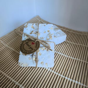 Puede incluir: Dos pastillas de jabón rectangulares de color blanco con copos de avena de color marrón claro. Las pastillas están atadas con cuerda marrón y tienen una etiqueta redonda de papel marrón con un corazón rojo y el texto "thank you" en negro.