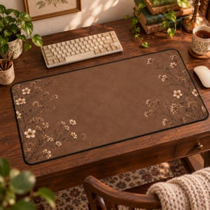 May include: A rectangular desk mat featuring a solid brown background with delicate, light-coloured floral branches extending from the left and right sides. The mat has a black stitched border and a smooth surface, designed to sit under a keyboard.