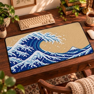 Peut inclure: Un tapis de bureau rectangulaire arborant un motif en pixel art représentant une grande vague bleue avec des crêtes d'écume blanche sur un fond beige. Le style graphique utilise des carrés pour illustrer l'eau et un petit sommet montagneux.