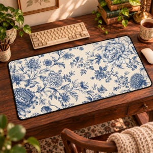 May include: A rectangular desk mat featuring a cream-colored background with a repeating blue floral pattern of chrysanthemums and various blossoms. The mat has a black stitched border and is positioned on a dark wooden desk surface.