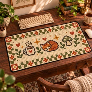 May include: A rectangular desk mat featuring a pixel art design on a cream background. The pattern includes a sleeping orange cat, a steaming mug with a heart, a mushroom, and small stars, all framed by a green and cream checkered border with flowers.