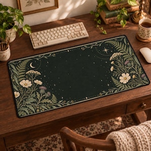 May include: A rectangular desk mat featuring a dark green background with a botanical illustration of white cosmos flowers, purple thistles, and green ferns. The design includes a crescent moon and small stars, creating a celestial theme.