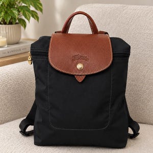 May include: A black backpack featuring a brown leather flap with a gold-tone snap button closure and a top handle. The bag has a zippered main compartment, visible stitching, and adjustable black shoulder straps, showcasing a structured design.