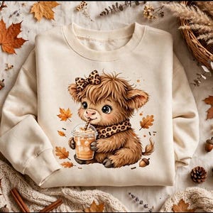 Peut inclure: Un sweat-shirt à col rond couleur crème orné d'une illustration graphique d'un veau Highland brun et duveteux portant un nœud et une écharpe à motif léopard, tenant un gobelet en plastique avec une paille, entouré de feuilles d'automne.