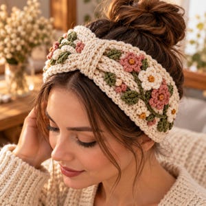 Op de afbeelding: Een crèmekleurige gehaakte hoofdband met een centraal gedraaid knoopontwerp. Het accessoire is versierd met kleine, driedimensionale bloemmotieven in roze en witte tinten, geaccentueerd door groene gehaakte blaadjes en steeltjes.