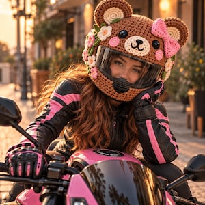 Puede incluir: Una funda de ganchillo marrón para casco con forma de oso, que incluye un lazo rosa, flores tejidas en blanco y rosa, y una visera transparente. La funda presenta orejas de oso redondeadas y un detalle de hocico, sobre un casco de moto.