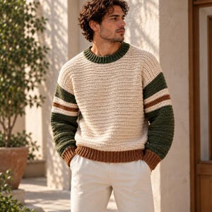 May include: A long-sleeved crochet jumper featuring a cream-coloured body with dark green and brown striped sleeves. The garment has a dark green ribbed crew neckline and a brown ribbed hem. The jumper is paired with plain white trousers.
