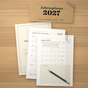 May include: A 2027 annual planner printable PDF set featuring a calendar page for January 2027, a yearly overview page, and a document with the text Oma. A silver paperclip attaches the pages, and a green pencil rests on the bottom sheet.