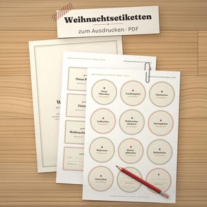 May include: A set of printable PDF sheets labeled "Weihnachtsetiketten zum Ausdrucken - PDF" featuring various circular and rectangular labels with German text, a silver paperclip, and a red pencil resting on the top sheet.