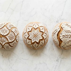 May include: Three round loaves of sourdough bread arranged in a horizontal row. Each loaf features a distinct, intricate scored pattern on its golden-brown crust, dusted with white flour, showcasing fan, floral, and wheat-like designs.
