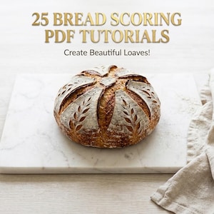 May include: A round, golden-brown sourdough loaf with intricate wheat-stalk scoring patterns on its crust. The text above reads 25 BREAD SCORING PDF TUTORIALS and Create Beautiful Loaves! The bread sits on a white marble board.