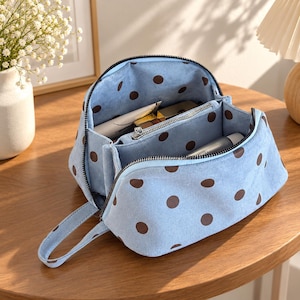 May include: A light blue cosmetic bag with a brown polka dot pattern, featuring a black zip closure, a side carrying strap, and an open interior compartment with a central zipped divider containing various small personal care items.