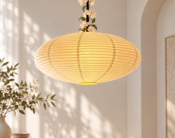 Pode incluir: Uma luminária pendente de papel em formato de lanterna, de cor creme e com nervuras horizontais. A luminária está suspensa por um fio escuro adornado com flores de cerejeira artificiais brancas que se estendem a partir do topo da cúpula.