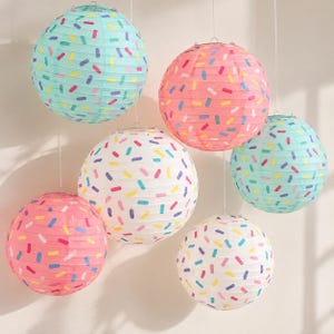 May include: A collection of six spherical paper lanterns hanging at varying heights. The lanterns feature a repeating pattern of small, colorful rectangular sprinkles in shades of pink, blue, yellow, and purple against white, light blue, and pink backgrounds.