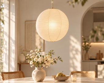 Puede incluir: Una lámpara colgante esférica con pantalla de papel blanco y finas nervaduras horizontales, suspendida por una delgada varilla metálica. La luz emite un brillo cálido y suave, iluminando el espacio circundante con una luz difusa.