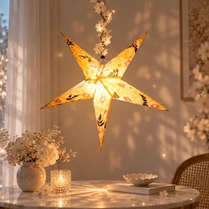 May include: A five-pointed star-shaped paper lantern with a warm yellow glow, featuring embedded pressed flower petals and dark green leaf patterns. The lantern hangs from a string adorned with small white blossoms and tiny fairy lights.