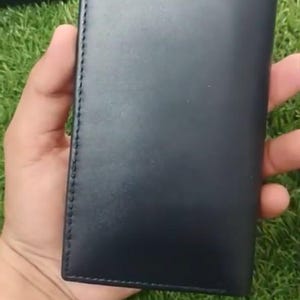 May include: A rectangular, dark navy blue leather wallet featuring a smooth, matte finish and visible tonal stitching along the vertical edge. The wallet is slim and elongated in shape, with a clean, unadorned surface and a structured, folded design.