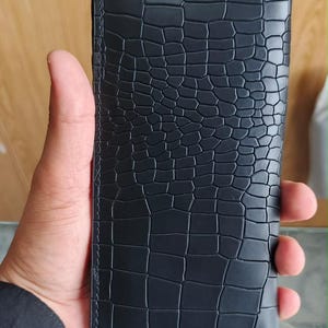 May include: A rectangular black wallet featuring a textured crocodile skin pattern across the entire surface. The wallet has straight edges and visible stitching along the perimeter, presenting a uniform and consistent dark finish throughout.