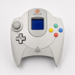 May include: A light gray Dreamcast video game controller featuring a central memory card slot with a blue screen, a circular analog stick, a directional pad, a Start button, and four colored action buttons labeled A, B, X, and Y on the right side.