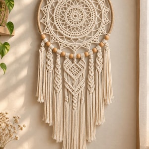 May include: A circular wall hanging featuring intricate off-white macrame cord knotted into a geometric mandala pattern. The design includes a central starburst, wooden beads along the lower arc, and long, flowing fringe with braided accents.