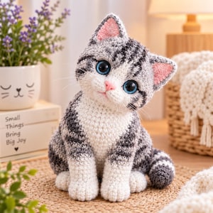 Op de afbeelding: Een gehaakt kattenfiguurtje met grijs en wit gestreepte vacht, grote blauwe ogen en een roze neus. De kat heeft puntige oren met roze details aan de binnenkant en zit in een rechtopstaande houding. De textuur van het garen is overal zichtbaar.