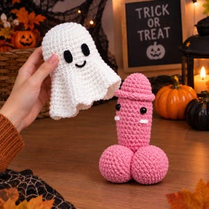 May include: A white crocheted ghost figure with black eyes and a smile, and a pink crocheted phallic-shaped figure with black eyes and white accents. The ghost is held above the pink figure, which stands upright on a wooden surface.
