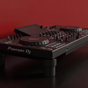 Op de afbeelding: Een zwarte Pioneer DJ-controller met twee ronde jogwheels, een centrale mixersectie met diverse knoppen en faders, een display aan de bovenzijde en verschillende performance-pads, geplaatst op vier cilindervormige zwarte steunvoeten.